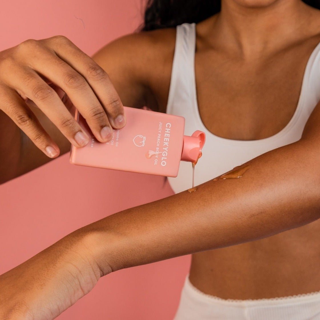 A person with long hair is applying CheekyGlo Juicy Peach Body Oil from a pink tube labeled &quot;CheekyGlo&quot; onto their forearm. They are wearing a white top, and the background is a solid pink color.