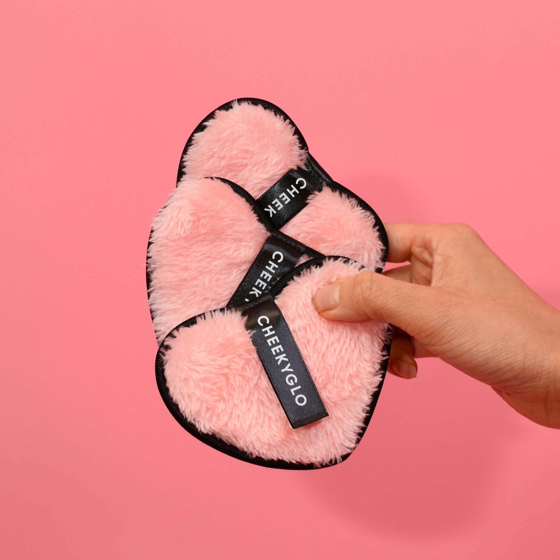 A hand is holding three pink, heart-shaped, fluffy CheekyGlo Makeup Erasers against a plain pink background. Each makeup eraser has a black tag with the brand name &quot;CheekyGlo&quot; in white letters. These CheekyGlo Makeup Erasers are perfect for eco-friendly skincare and serve as reusable makeup removers.