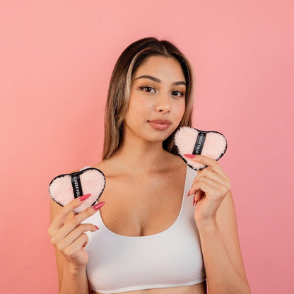 A woman with long hair stands against a pink background, holding two heart-shaped, pink fluffy pads edged in black with tags in each hand. She is wearing a white tank top and looking directly at the camera. These CheekyGlo Makeup Erasers by CheekyGlo are perfect for eco-friendly skincare lovers seeking reusable makeup removers.