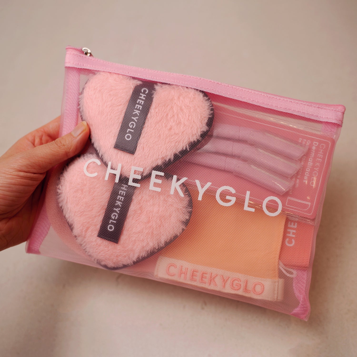 A hand holds a chic CheekyGlo Reward 2 pink mesh pouch featuring fluffy pink heart-shaped items, gloves, and a folded bamboo silk eye mask. The neutral backdrop enhances the elegant appeal of this makeup bag from CheekyGlo.