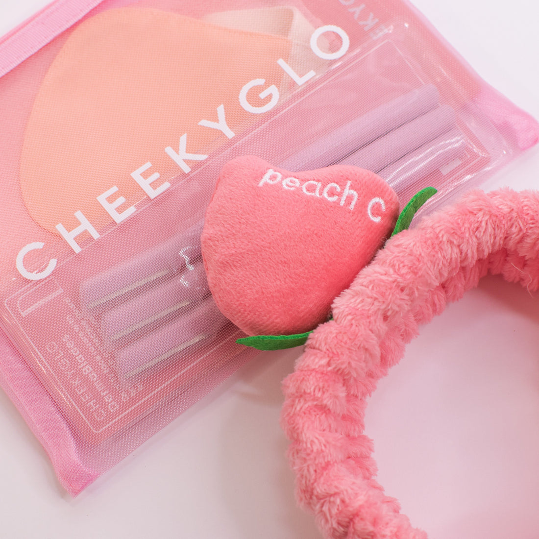 The CheekyGlo Peachy Headband, featuring a plush peach accessory with the word "peach" and the letter "C" embroidered on it, sits next to a pink CHEEKYGLO pouch filled with other items. This fluffy elastic headband is ideal for your self-care routine and looks stunning against a light background.