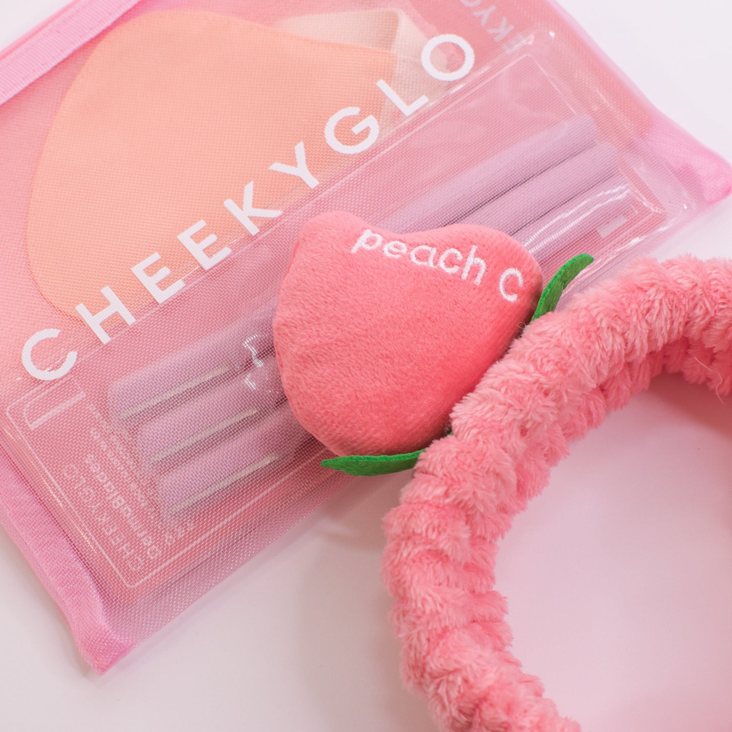 The CheekyGlo Peachy Headband, featuring a plush peach accessory with the word &quot;peach&quot; and the letter &quot;C&quot; embroidered on it, sits next to a pink CHEEKYGLO pouch filled with other items. This fluffy elastic headband is ideal for your self-care routine and looks stunning against a light background.