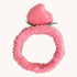 Introducing the CheekyGlo Peachy Headband by CheekyGlo: a pink plush scrunchie featuring a decorative peach on top with "peachc" embroidered in white text. This fluffy elastic headband elevates your self-care routine, providing both style and comfort with its soft and fluffy material.