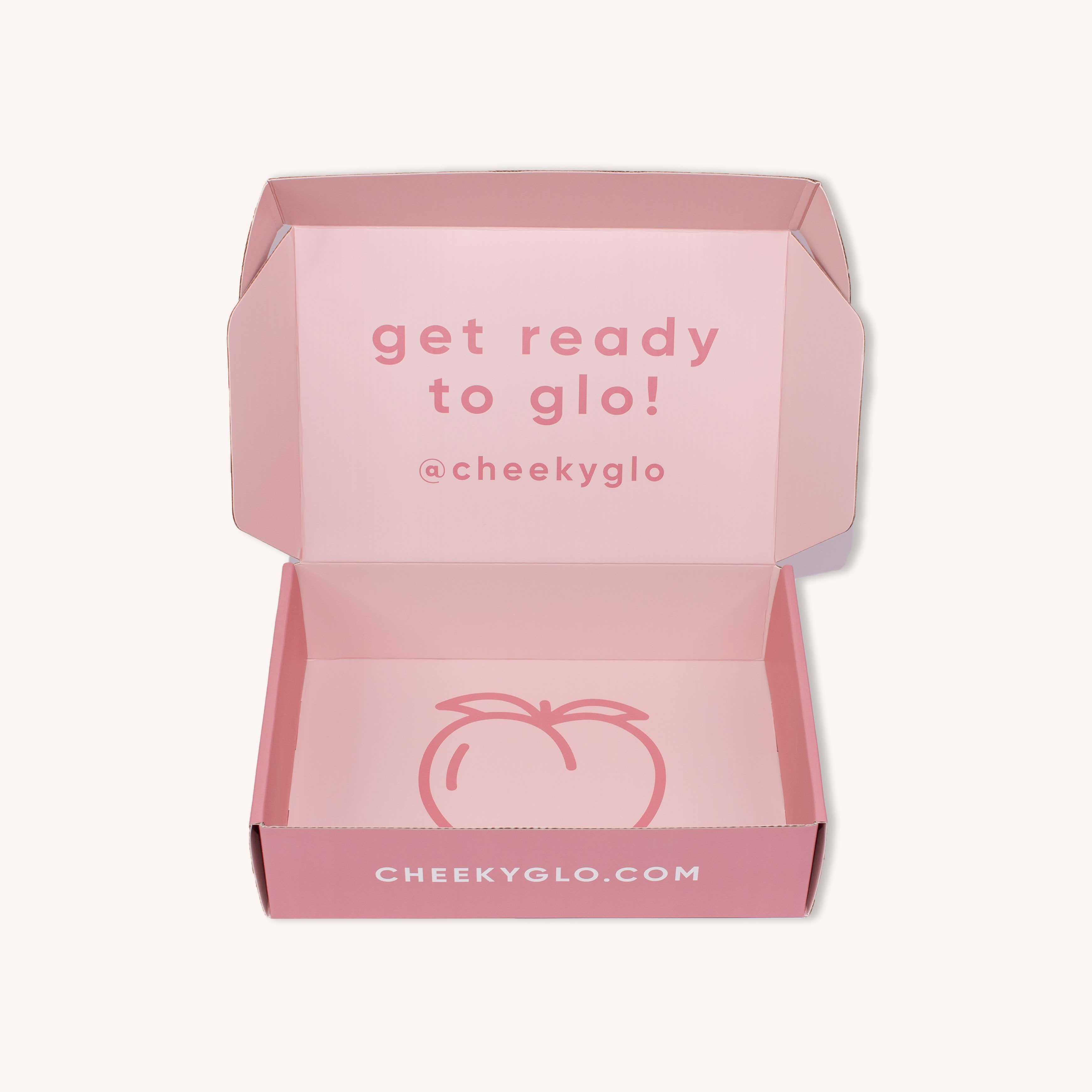 A CheekyGlo Premium Gift Box in pink cardboard with the lid open, revealing the inside. The inner lid reads &quot;get ready to glo!&quot; with &quot;@cheekyglo&quot; underneath. This premium self-care gift box from CheekyGlo displays the website &quot;CHEEKYGLO.COM&quot; and features an illustration of a peach at the bottom part of the box.