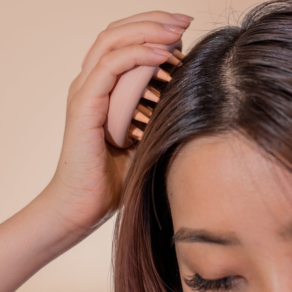 Close-up of a person using the CheekyGlo Scalp Scrubber on their hair. The individual's hand is holding the CheekyGlo scrubber against their scalp, moving it through their hair for healthy hair growth. The background is beige, and the person's face is partially visible, showing their eye and eyebrow.