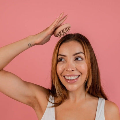 A woman with long brown hair is smiling while using a CheekyGlo Scalp Scrubber on her head. She is wearing a white sleeveless top and standing against a pink background, promoting healthy hair growth for the CheekyGlo brand.