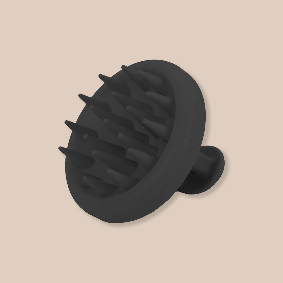 The CheekyGlo Scalp Scrubber by CheekyGlo, featuring soft, flexible black silicone bristles and a round handle, rests against a beige background. This tool is perfect for promoting healthy hair growth and serves as an effective scalp scrubber.