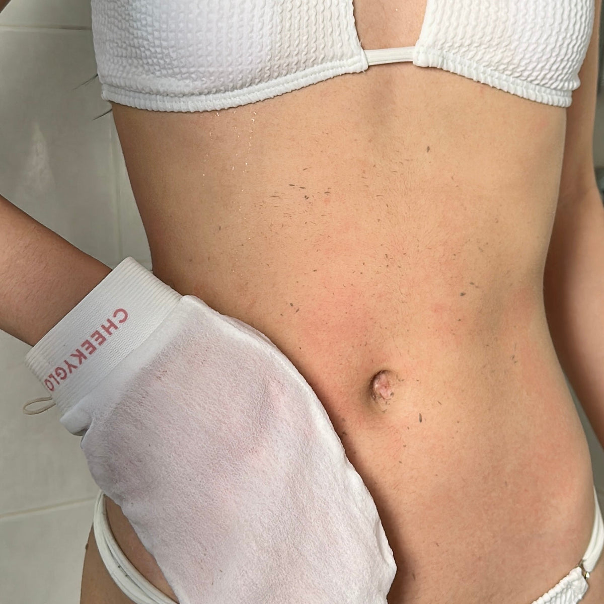 Close-up of a person's midsection wearing a white bikini, holding a CheekyGlo Sensitive Exfoliating Glove. They appear to be scrubbing their skin with this eczema-friendly exfoliator against the backdrop of a plain bathroom wall.