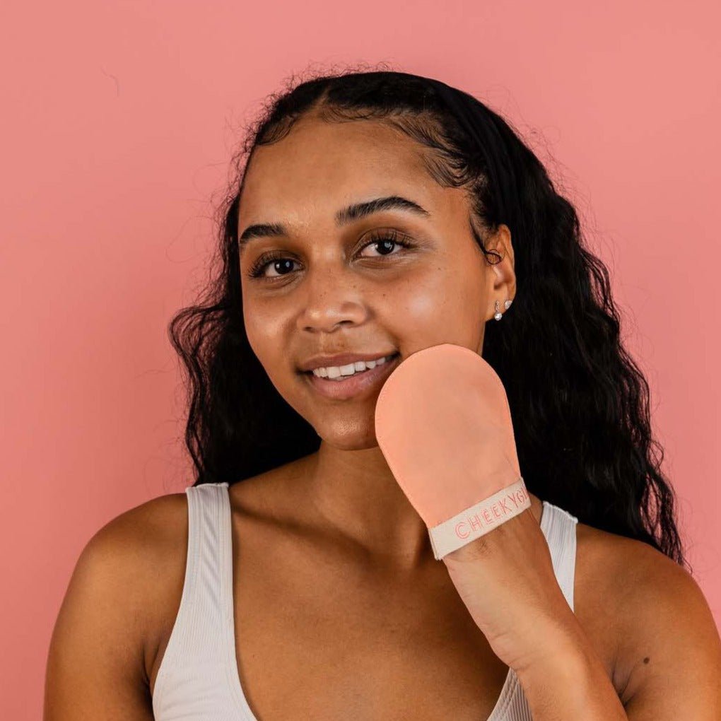 A smiling person with long, curly hair is holding a CheekyGlo Silk Exfoliating Face Mitt made from premium Turkish silk, featuring the word &quot;CHEEKYS.&quot; They press it against their face while standing against a solid pink background and wearing a light-colored tank top.