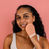 A smiling person with long, curly hair is holding a CheekyGlo Silk Exfoliating Face Mitt made from premium Turkish silk, featuring the word "CHEEKYS." They press it against their face while standing against a solid pink background and wearing a light-colored tank top.
