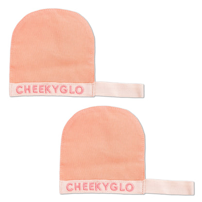 Two peach-colored CheekyGlo Silk Exfoliating Face Mitts, crafted from premium Turkish silk, with &quot;CHEEKYGLO&quot; embroidered in pink on the white wrist straps. The mitts are displayed side by side, slightly overlapping, against a white background.