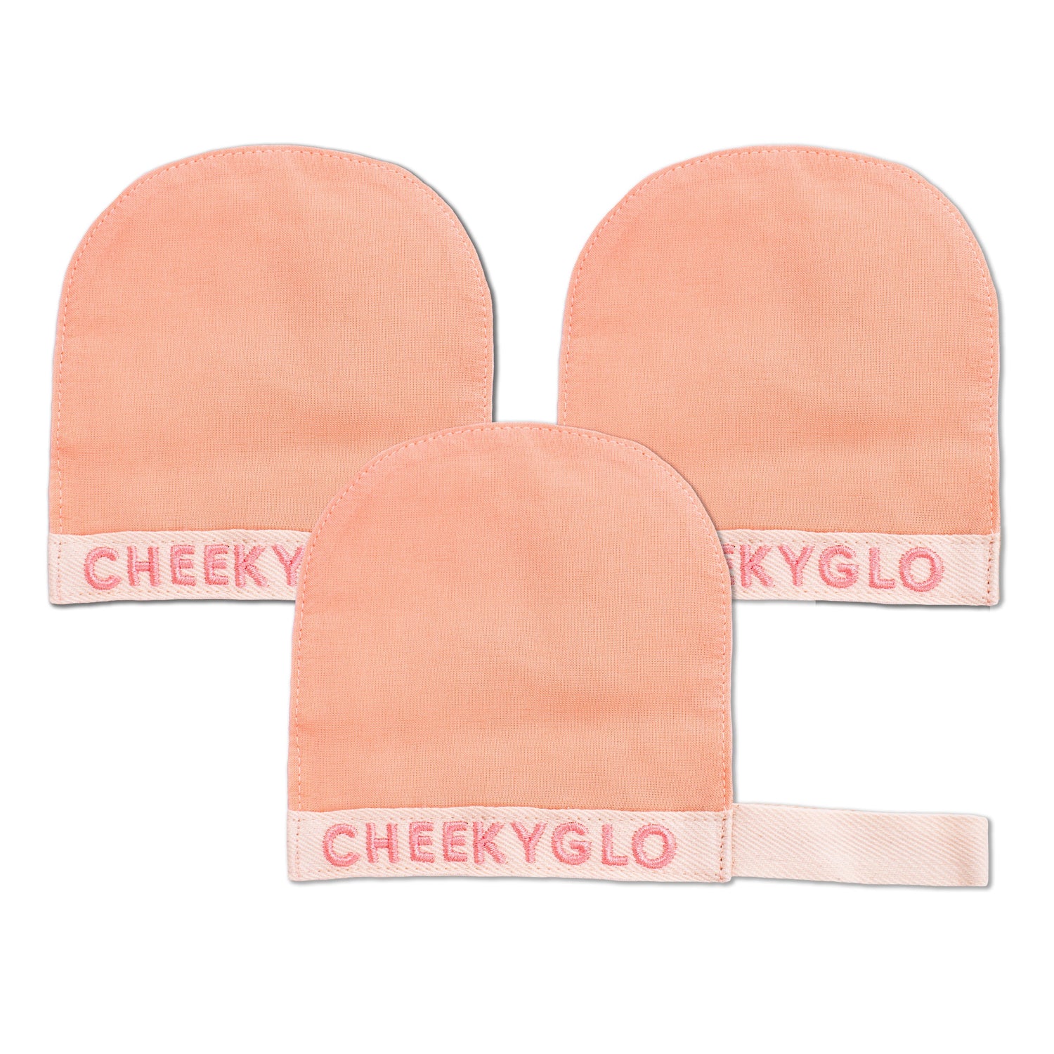 A triangular arrangement of three peach-colored CheekyGlo Silk Exfoliating Face Mitts, crafted from Premium Turkish Silk, is displayed on a white background. Each mitt features &quot;CheekyGlo&quot; embroidered in pink on a white band at the bottom, and one mitt includes a looped strap for convenient hanging.