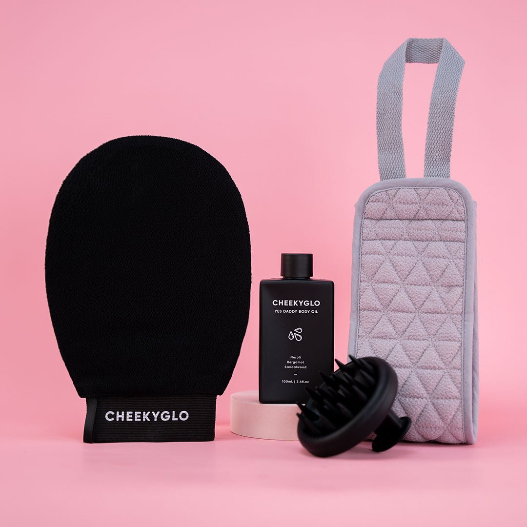 The Everyday Essentials Set by CheekyGlo, displayed on a pink background, includes an exfoliating body glove, a bottle of body gel, a gray textured carrying case, a scalp scrubber massager, and a roll of tape for your beauty routine.