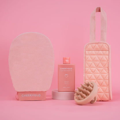 Introducing the CheekyGlo Everyday Essentials Set: a pink-themed skincare collection that includes a textured exfoliating body glove, a bottle of body oil, a quilted pouch, and a scalp massager, beautifully presented against a pink background.