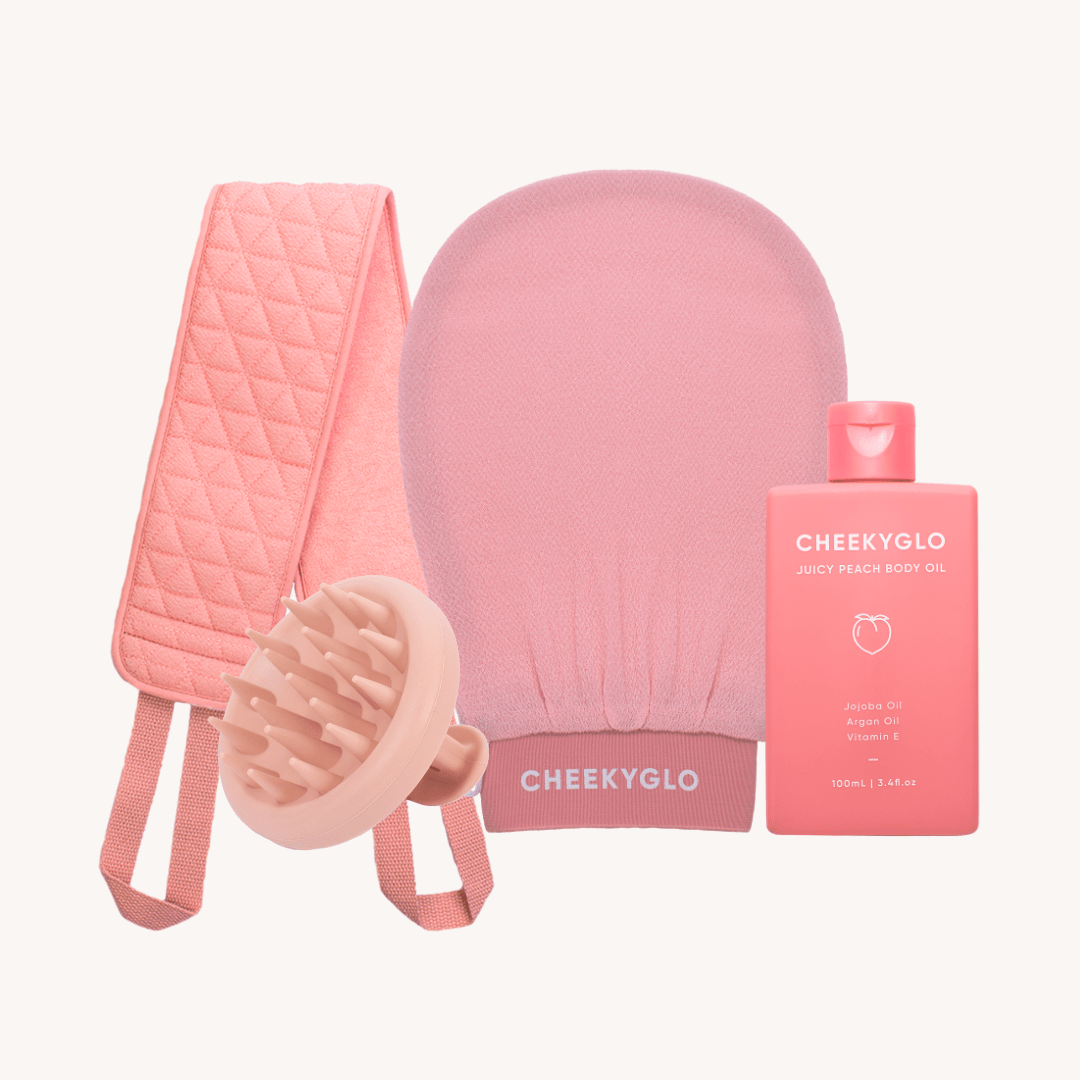 The Everyday Essentials Set by CheekyGlo includes an assortment of pink skincare products, such as an exfoliating body glove, a quilted strap, and a revitalizing scalp scrubber. Additionally, it features a bottle of CheekyGlo Juicy Peach Body Oil. These items are elegantly displayed against a light backdrop.