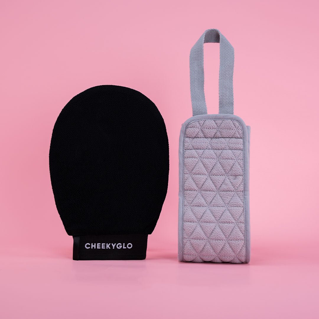 The Exfoliating Body Duo by CheekyGlo includes a black body glove crafted from biodegradable viscose, complemented by a gray textured pouch with a handle, all showcased against a vibrant pink background.