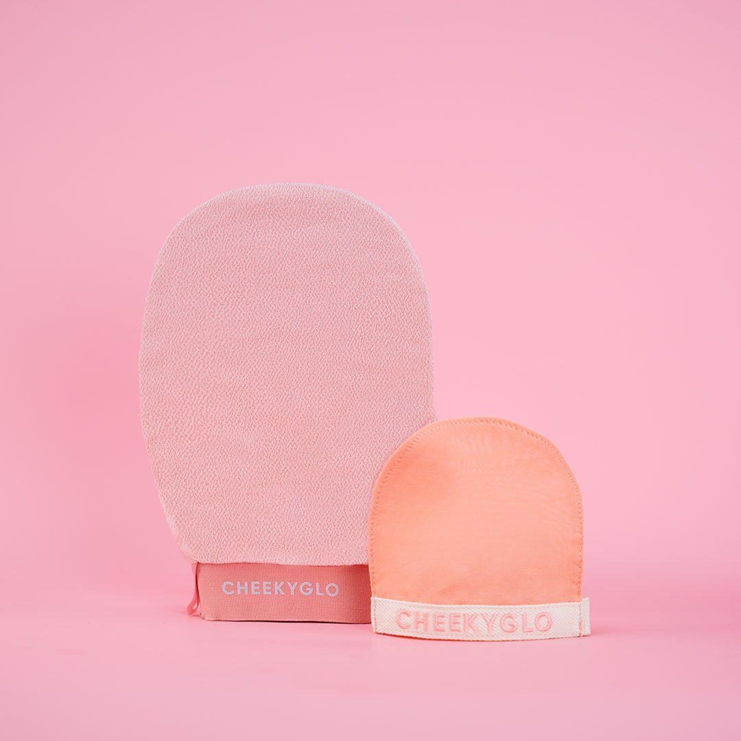 A light pink CheekyGlo Face+Body Exfoliating Duo and a smaller peach-colored glove, both labeled &quot;CheekyGlo,&quot; are displayed against a pink background. The larger mitt is standing upright, while the smaller one is positioned in front of it.