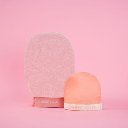 A light pink CheekyGlo Face+Body Exfoliating Duo and a smaller peach-colored glove, both labeled &quot;CheekyGlo,&quot; are displayed against a pink background. The larger mitt is standing upright, while the smaller one is positioned in front of it.