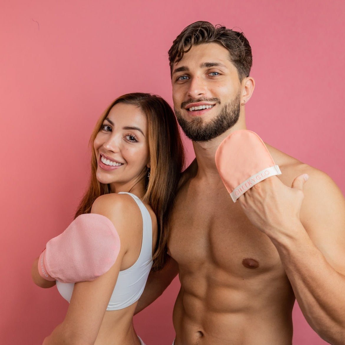 A smiling man and woman pose against a pink background, both holding CheekyGlo Face+Body Exfoliating Duo mitts. The woman, with long brown hair in a white crop top, stands beside the shirtless man with short brown hair and a beard. Each has an exfoliating mitt on one hand, showcasing the Face+Body Exfoliating Duo product.