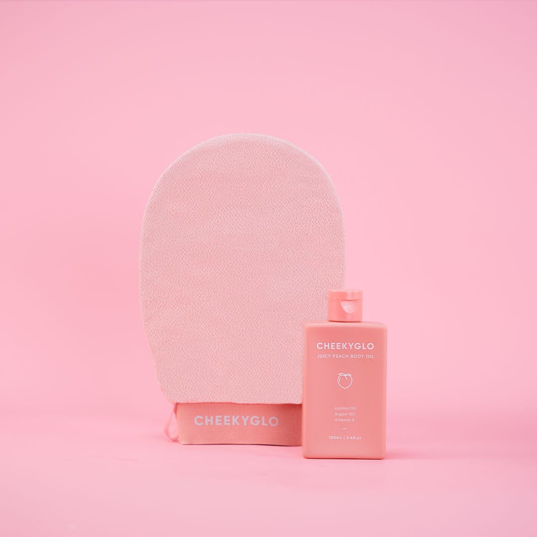 The image features the CheekyGlo Glow Up Duo [MOST POPULAR], showcasing a light pink exfoliating glove standing upright next to its matching peachy-pink rectangular bottle of body oil for sensitive skin. Both items are placed on a solid pink background, with the bottle labeled &quot;CheekyGlo.