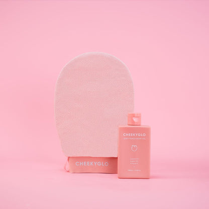 The image features the CheekyGlo Glow Up Duo [MOST POPULAR], showcasing a light pink exfoliating glove standing upright next to its matching peachy-pink rectangular bottle of body oil for sensitive skin. Both items are placed on a solid pink background, with the bottle labeled &quot;CheekyGlo.