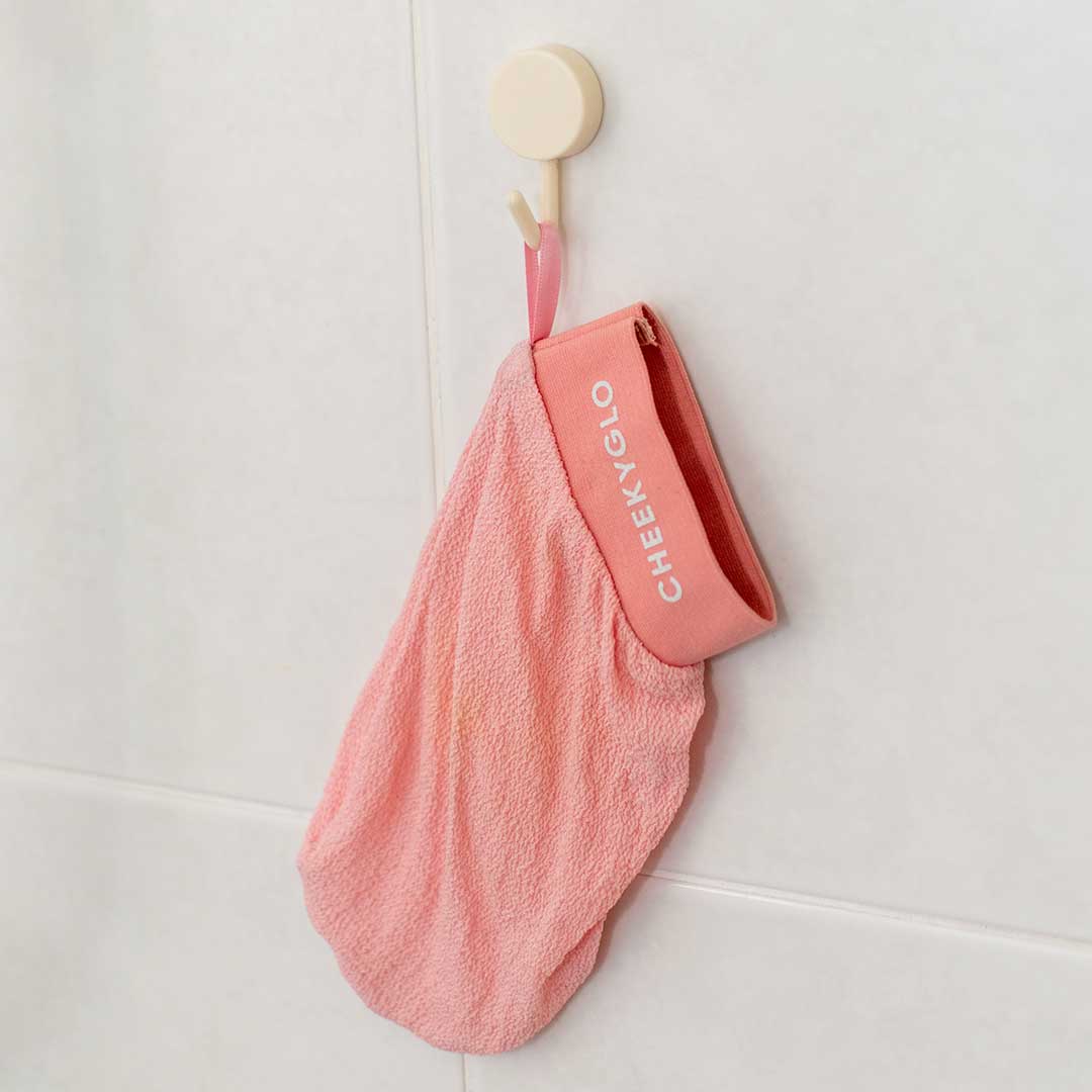 A pink exfoliating glove with a strap labeled &quot;CheekyGlo&quot; hangs from a rust-proof CheekyGlo Hanging Hook on a white tiled wall.