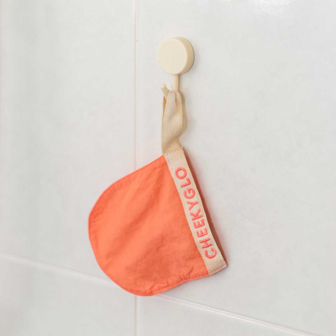 A CheekyGlo coral-colored fabric mitt with a beige strap hangs on a rust-proof, round Hanging Hook against a white tile wall.