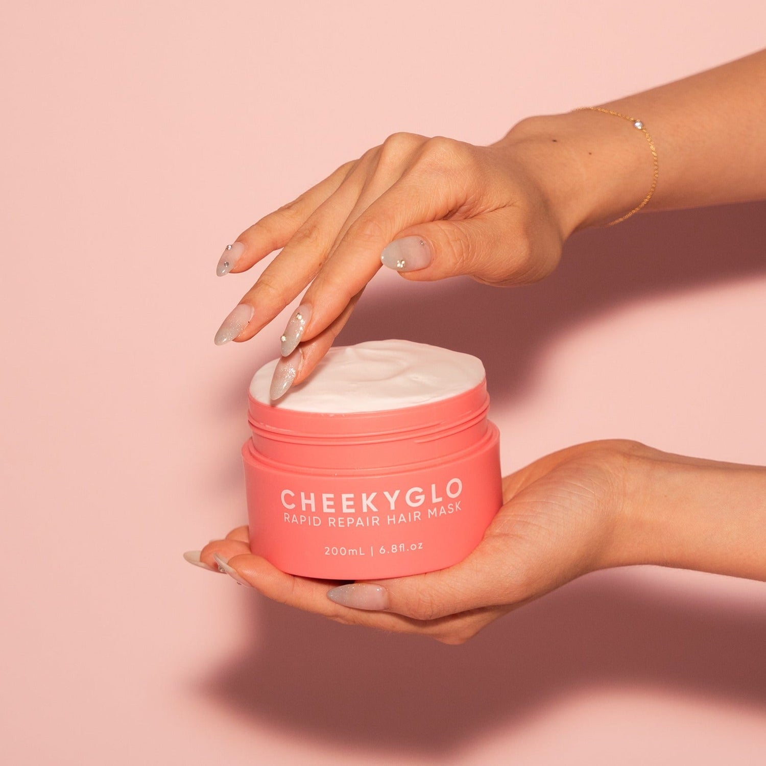 A person with manicured nails and an elegant bracelet is holding a pink jar of &quot;CheekyGlo Rapid Repair Hair Mask.&quot; The jar is open, revealing the creamy product inside, brimming with nourishing ingredients for smooth and shiny hair. The light pink background perfectly complements the jar&