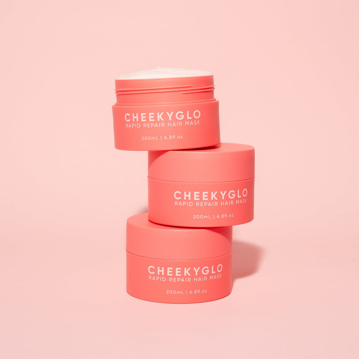 Three pink containers of CheekyGlo's Rapid Repair Hair Mask, packed with nourishing ingredients, are arranged in a pyramid shape against a soft pink background. The containers, marked with white text displaying the product name and size (200mL / 6.8 fl oz), promise smooth and shiny hair.
