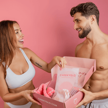 A woman and a man, both in white undergarments, smile at each other while holding a pink box filled with CheekyGlo&