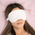 A person with long hair reclines on a plush, pink surface, wearing a CheekyGlo Anti-Acne Bamboo Silk Eye Mask. The image evokes relaxation and restfulness, further enhanced by the mask&